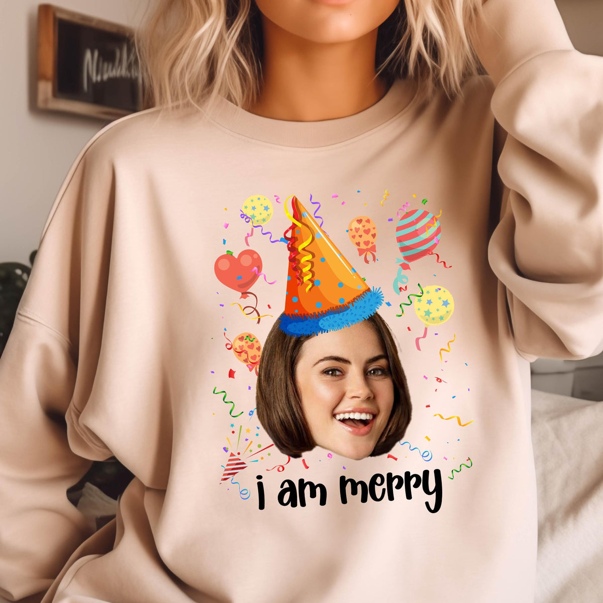 May include: A light pink sweatshirt with a colorful birthday party design. The design features a person's face with a party hat, confetti, and balloons. The text "i am merry" is printed below the design.
