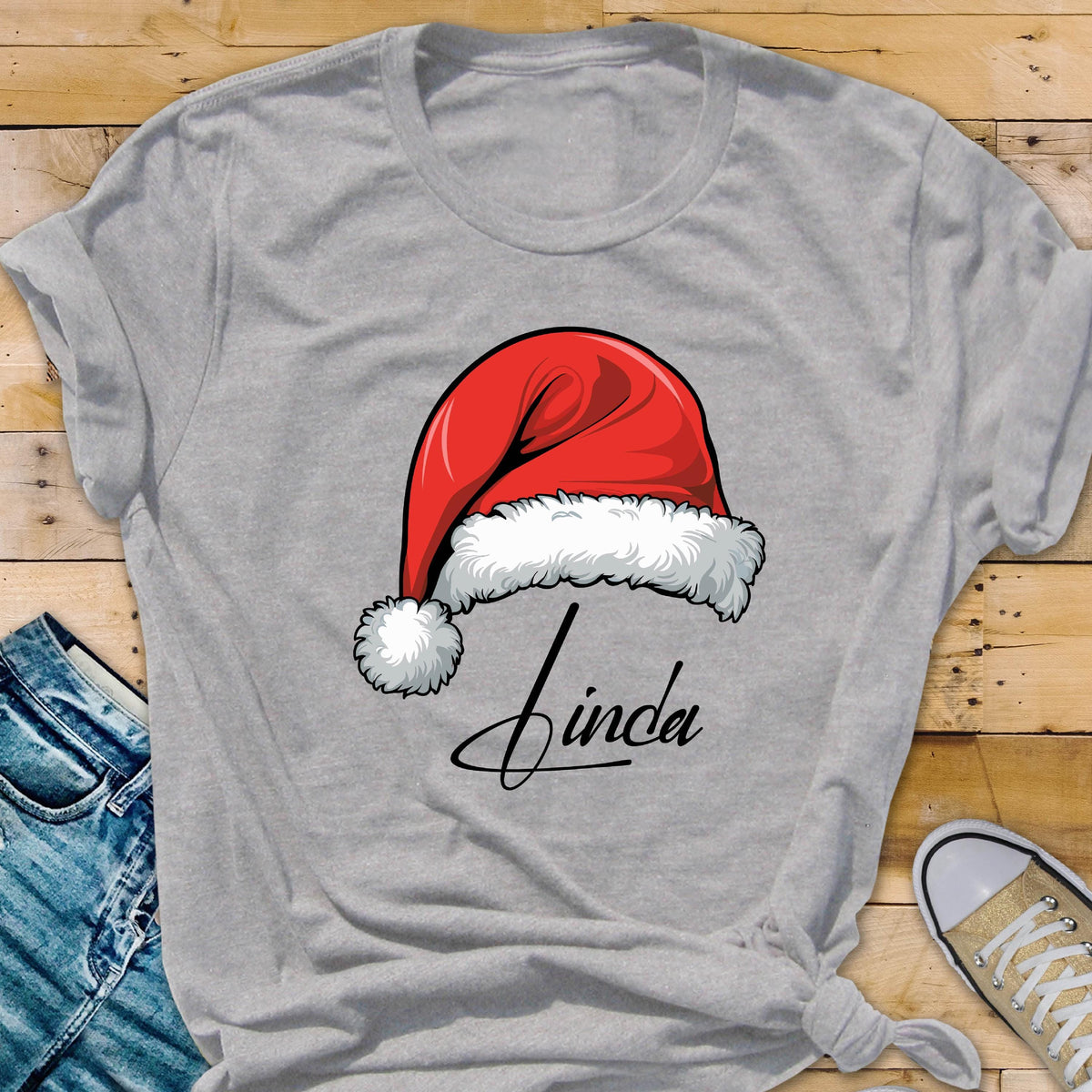 May include: A heather gray short-sleeved t-shirt featuring a graphic of a red Santa hat with white fur trim and a pom-pom. Below the hat, the name Linda is printed in a black cursive script font. The shirt has a crew neckline and short sleeves.