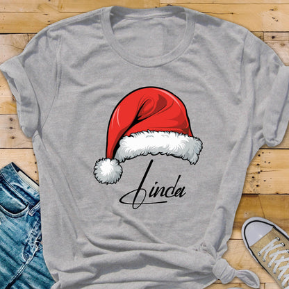May include: A heather gray short-sleeved t-shirt featuring a graphic of a red Santa hat with white fur trim and a pom-pom. Below the hat, the name Linda is printed in a black cursive script font. The shirt has a crew neckline and short sleeves.