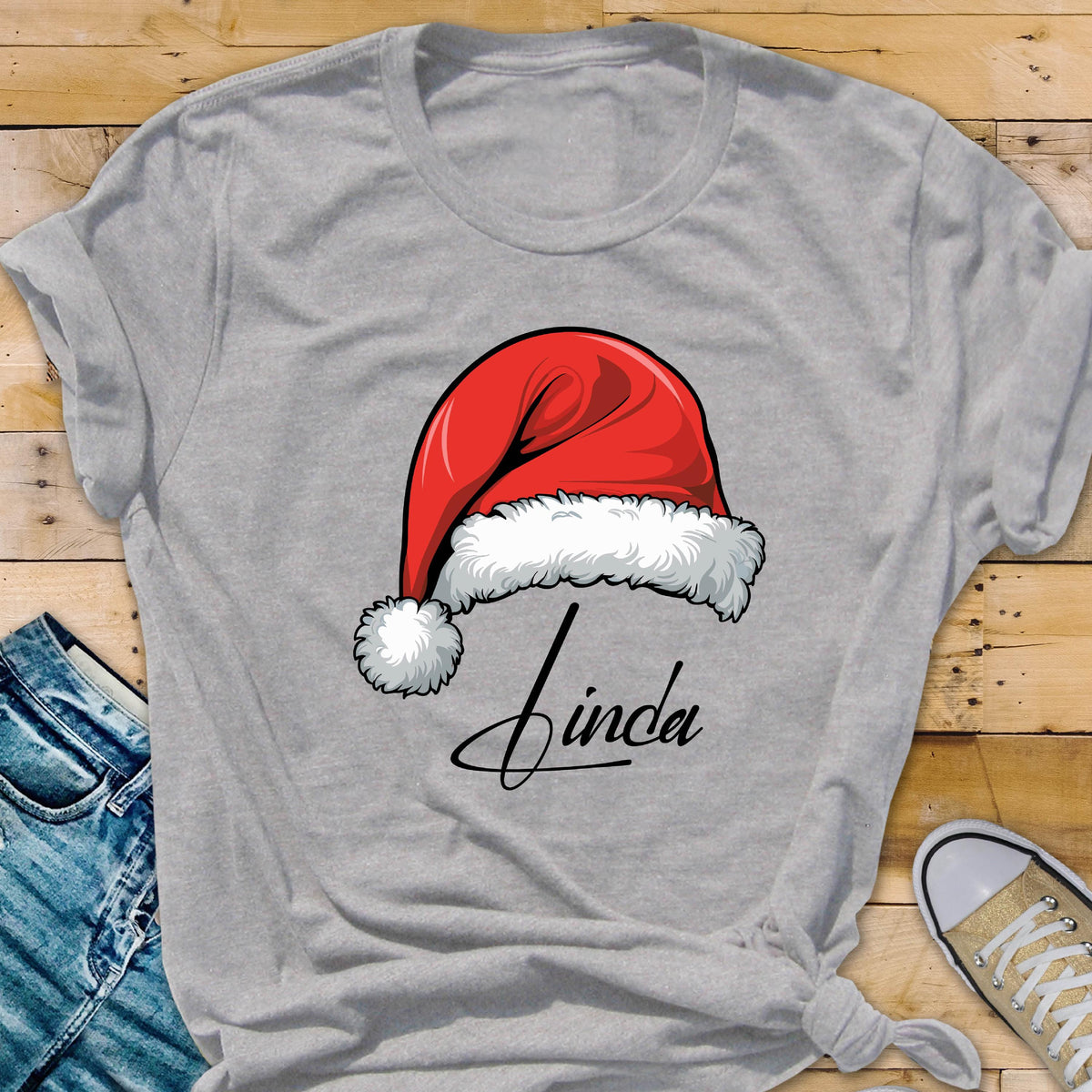 May include: A heather gray short-sleeved t-shirt featuring a graphic of a red Santa hat with white fur trim and a pom-pom. Below the hat, the name Linda is printed in a black cursive script font. The shirt has a crew neckline and short sleeves.