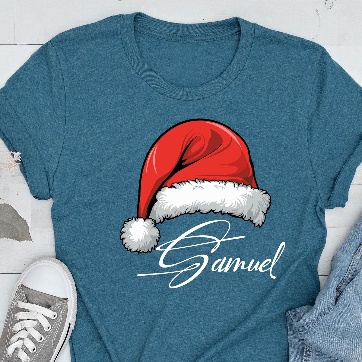 May include: A heathered teal short-sleeve t-shirt featuring a graphic of a red Santa hat with white fur trim and a pom-pom. Below the hat, the name Samuel is printed in a white, cursive script font. The shirt has a crew neckline.