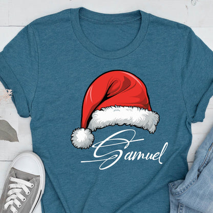 May include: A heathered teal short-sleeve t-shirt featuring a graphic of a red Santa hat with white fur trim and a pom-pom. Below the hat, the name Samuel is printed in a white, cursive script font. The shirt has a crew neckline.