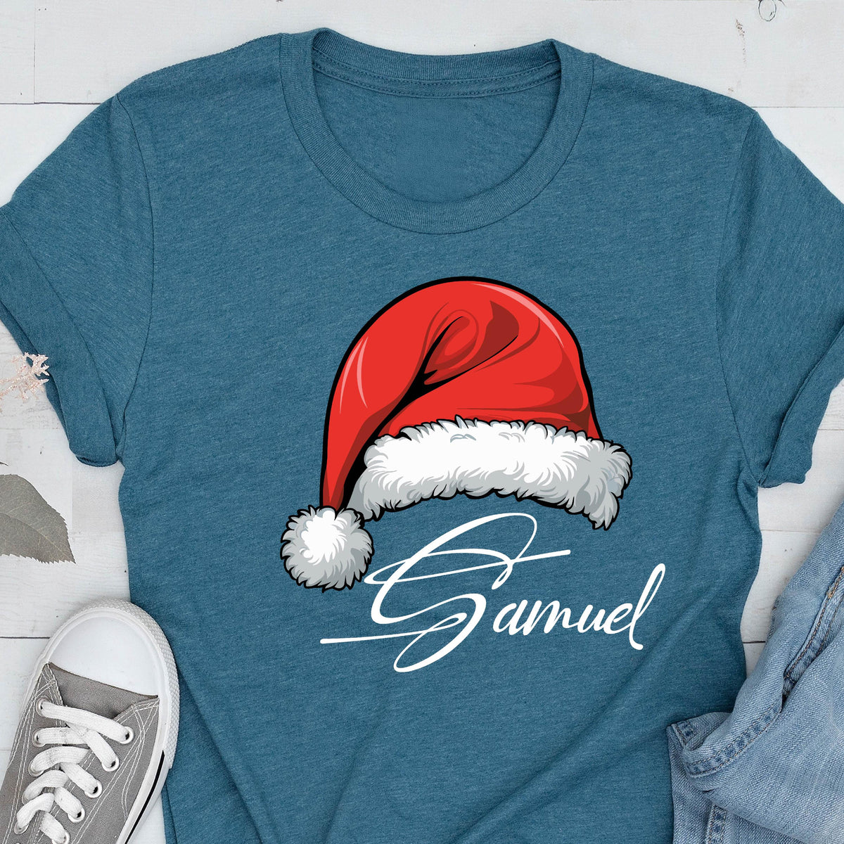 May include: A heathered teal short-sleeve t-shirt featuring a graphic of a red Santa hat with white fur trim and a pom-pom. Below the hat, the name Samuel is printed in a white, cursive script font. The shirt has a crew neckline.