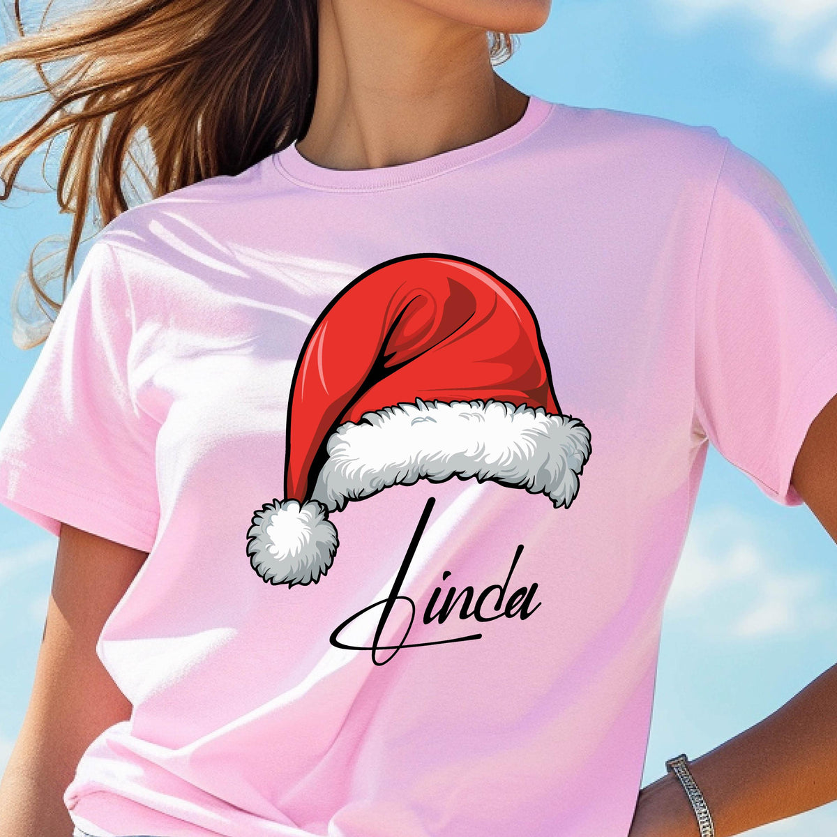 May include: A light pink short-sleeved t-shirt featuring a graphic of a red Santa hat with white fur trim and a pom-pom. Below the hat, the name Linda is printed in a black cursive script font. The shirt has a crew neckline and short sleeves.