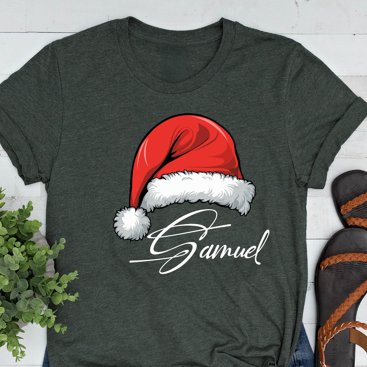 May include: A dark charcoal gray short-sleeve t-shirt featuring a graphic of a red Santa hat with a white fur trim and pom-pom. Below the hat, the name Samuel is printed in a white cursive script font across the center of the chest.