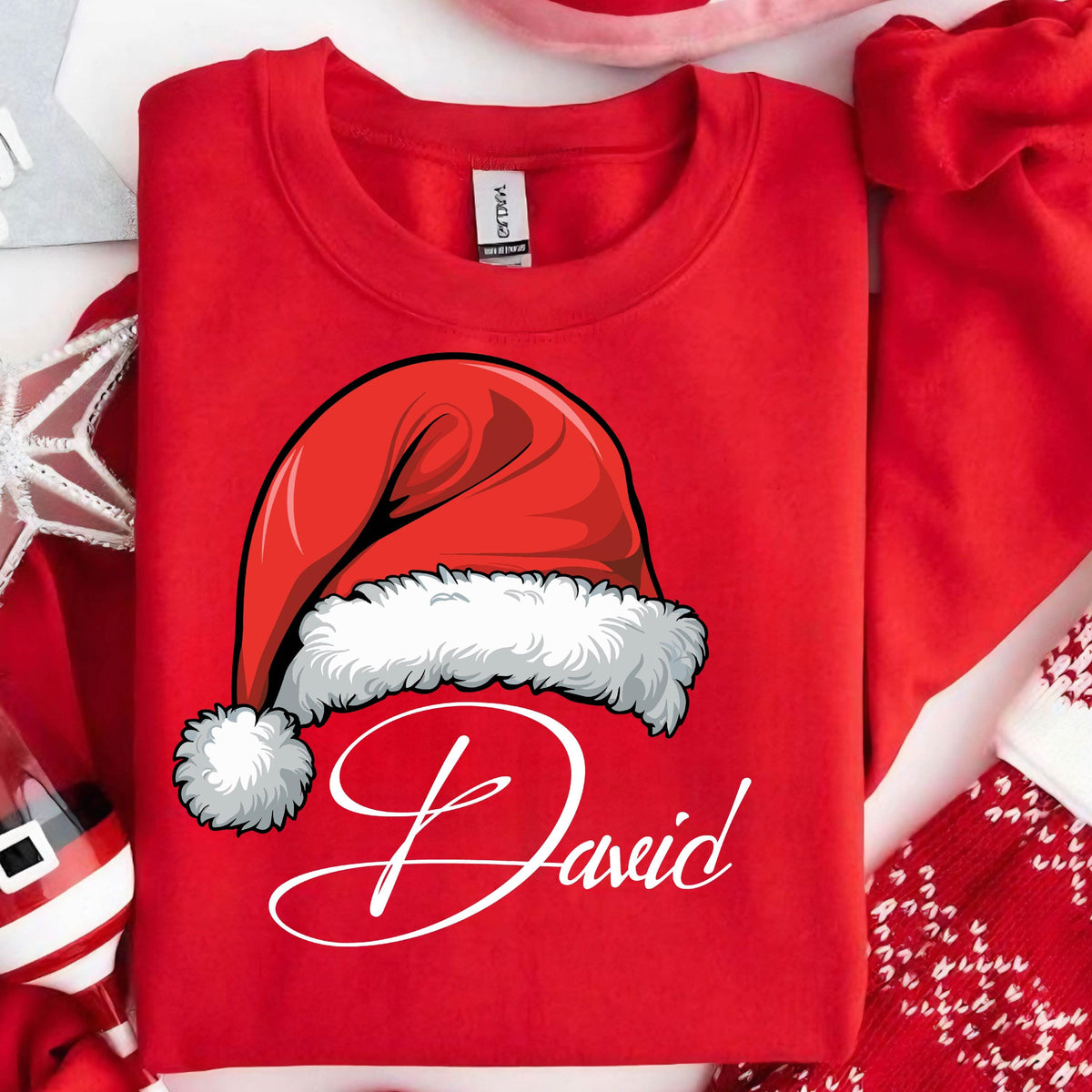 May include: A bright red crewneck sweatshirt featuring a graphic of a red Santa hat with a white fur trim and pom-pom. Below the hat, the name David is printed in a white cursive script font. The garment has a standard collar and long sleeves.