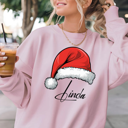 May include: A light pink long-sleeve sweatshirt featuring a graphic of a red and white Santa hat positioned above the name Linda written in black cursive script. The garment has a crew neckline and a relaxed fit, showcasing a festive holiday design.