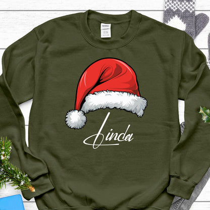 May include: An olive green crewneck sweatshirt featuring a graphic of a red Santa hat with a white fur trim and pom-pom. Below the hat, the name Linda is printed in a white cursive script font. The garment has long sleeves and a ribbed neckline.