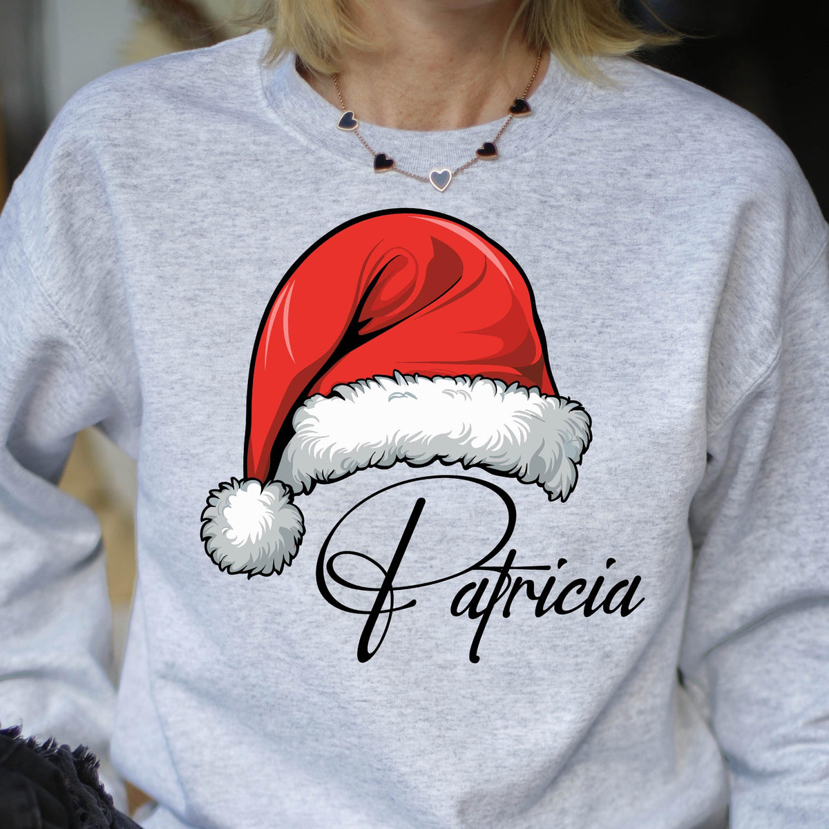 May include: A light gray crewneck sweatshirt featuring a graphic of a red Santa hat with a white fur trim and pom-pom. Below the hat, the name Patricia is printed in a black cursive script font. The garment has long sleeves and a ribbed neckline.