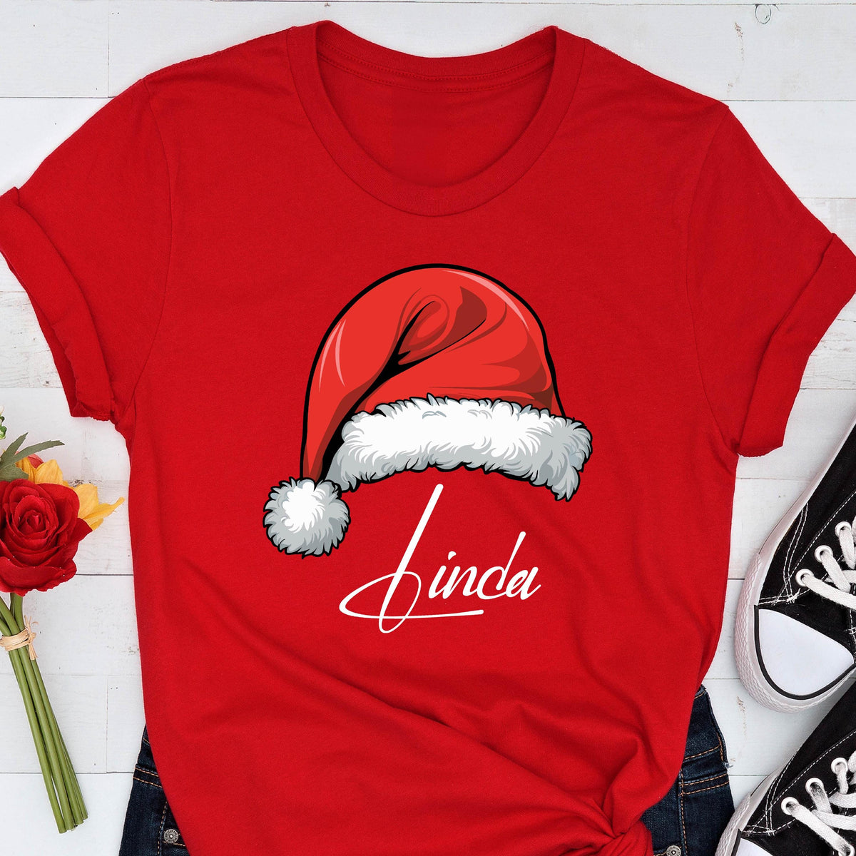 May include: A bright red short-sleeved t-shirt featuring a graphic of a red Santa hat with a white fur trim and pom-pom. Below the hat, the name Linda is printed in a white cursive script font. The shirt has a crew neckline and cuffed sleeves.
