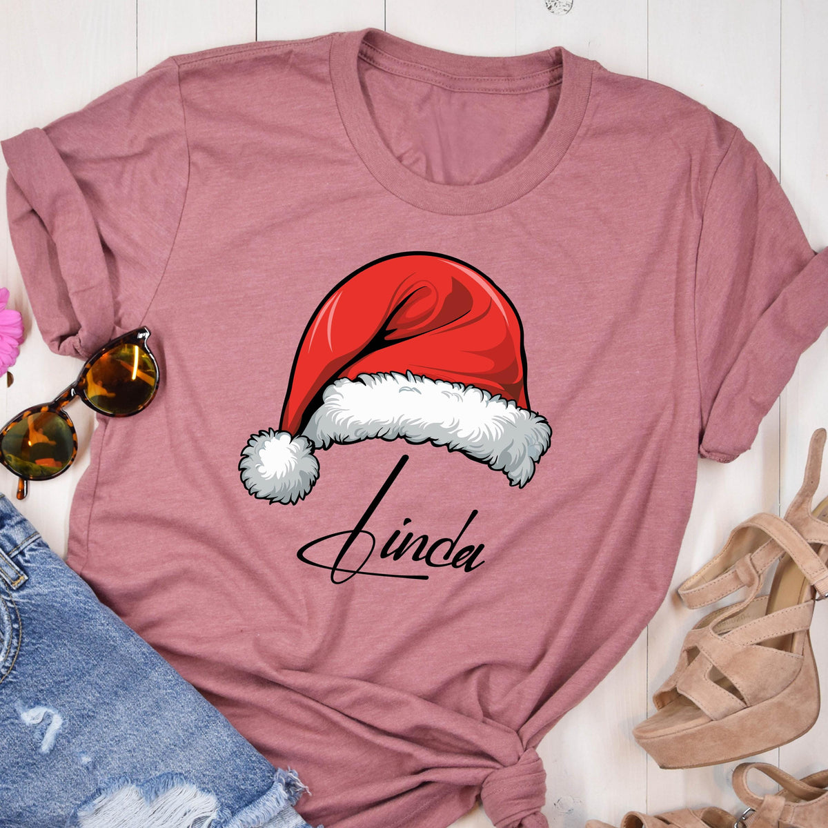 May include: A mauve short-sleeved t-shirt featuring a graphic of a red Santa hat with white fur trim and the name Linda written in black cursive script underneath the hat. The shirt has a crew neckline and a casual, relaxed fit design.