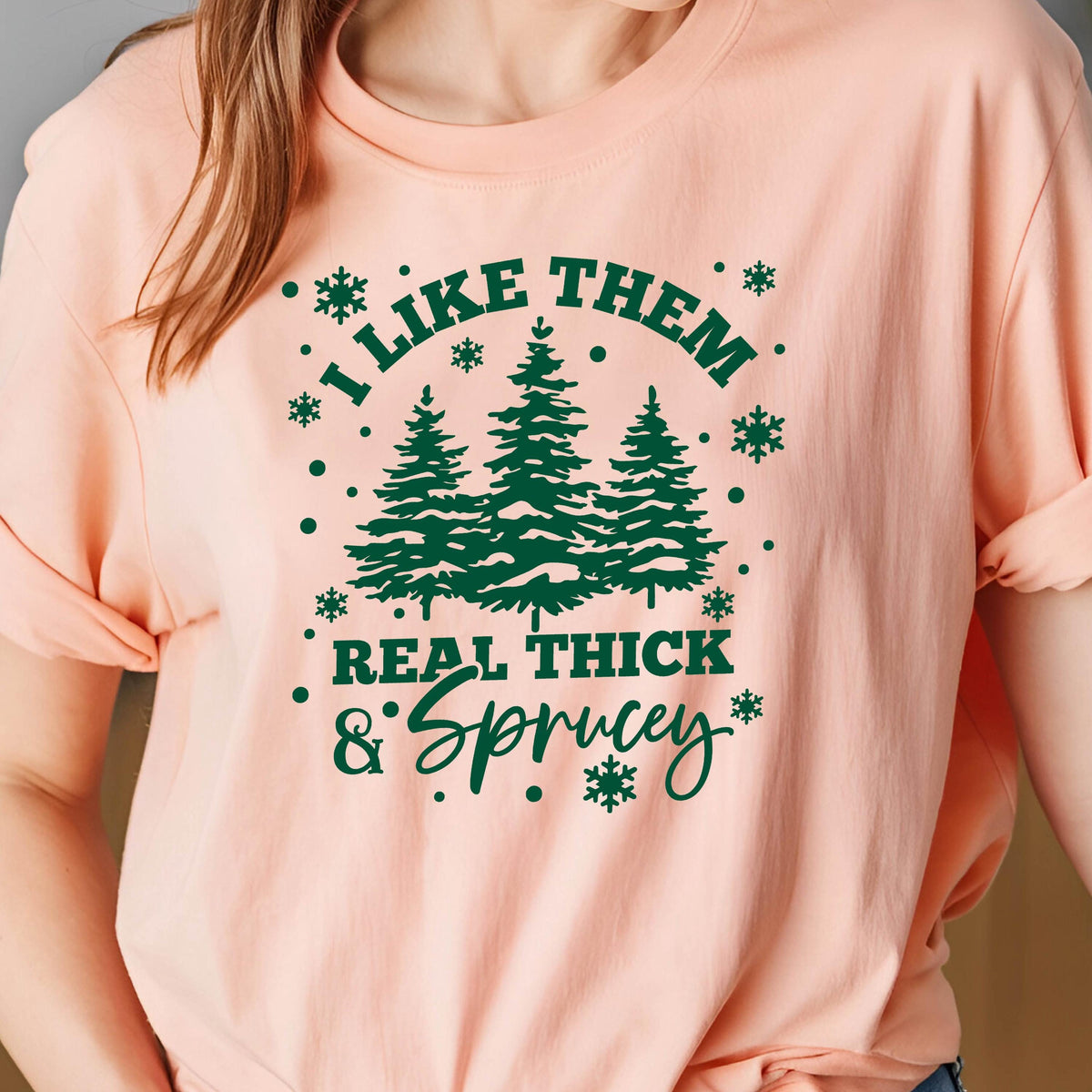May include: A light peach-colored t-shirt with the text "I LIKE THEM REAL THICK & Sprucey" in dark green. The design includes three green fir trees and snowflake accents.