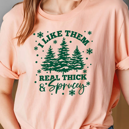 May include: A light peach-colored t-shirt with the text "I LIKE THEM REAL THICK & Sprucey" in dark green. The design includes three green fir trees and snowflake accents.