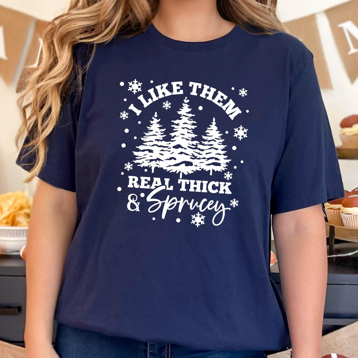 May include: Navy blue t-shirt with a white graphic featuring three stylized evergreen trees and the text "I LIKE THEM REAL THICK & SPRUCEY" in a whimsical font. Snowflakes and dots surround the text and trees.