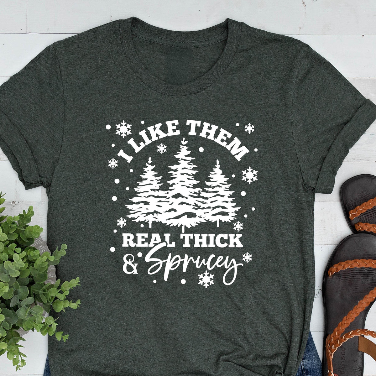 May include: Dark gray t-shirt with white text and graphics. The text reads "I LIKE THEM REAL THICK & SPRUCEY" with a graphic of three stylized evergreen trees. Snowflakes and dots surround the text.