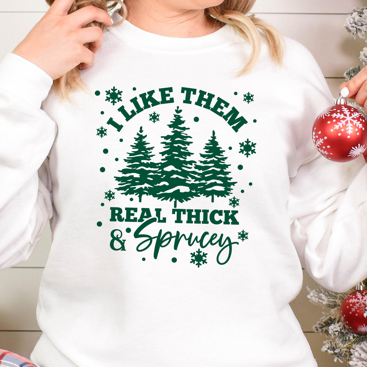 May include: A white long-sleeve sweatshirt featuring a dark green graphic print of three pine trees surrounded by snowflakes. The text reads I LIKE THEM REAL THICK & SPRUCEY in a mix of block and script fonts, centered on the chest area.