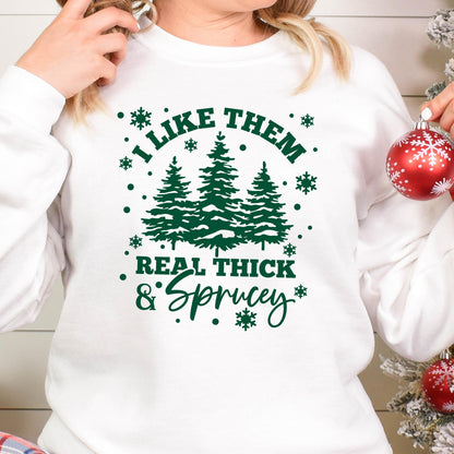 May include: A white long-sleeve sweatshirt featuring a dark green graphic print of three pine trees surrounded by snowflakes. The text reads I LIKE THEM REAL THICK & SPRUCEY in a mix of block and script fonts, centered on the chest area.