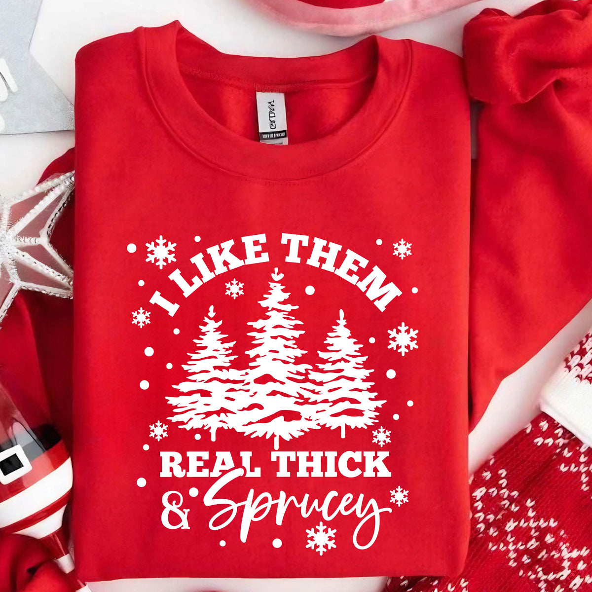 May include: A bright red crewneck sweatshirt featuring a white graphic print of three pine trees surrounded by snowflakes. The text reads, I LIKE THEM REAL THICK & SPRUCEY, arranged in a circular pattern around the central tree design.