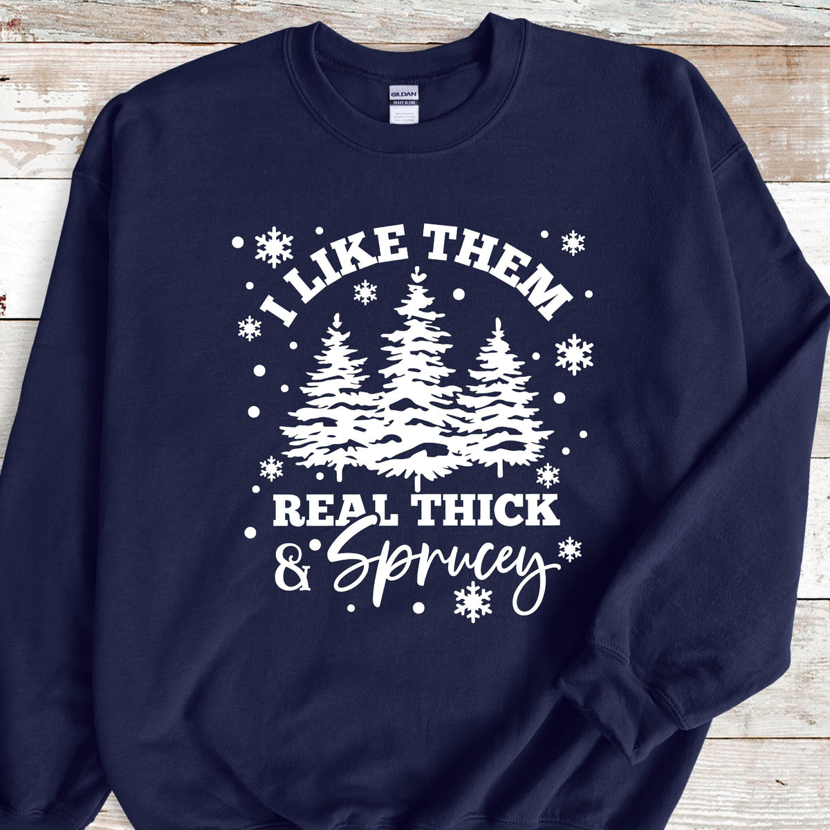 May include: A navy blue long-sleeve crewneck sweatshirt featuring a white graphic print of three pine trees surrounded by snowflakes. The text reads I LIKE THEM REAL THICK & SPRUCEY in a combination of bold block and script lettering.