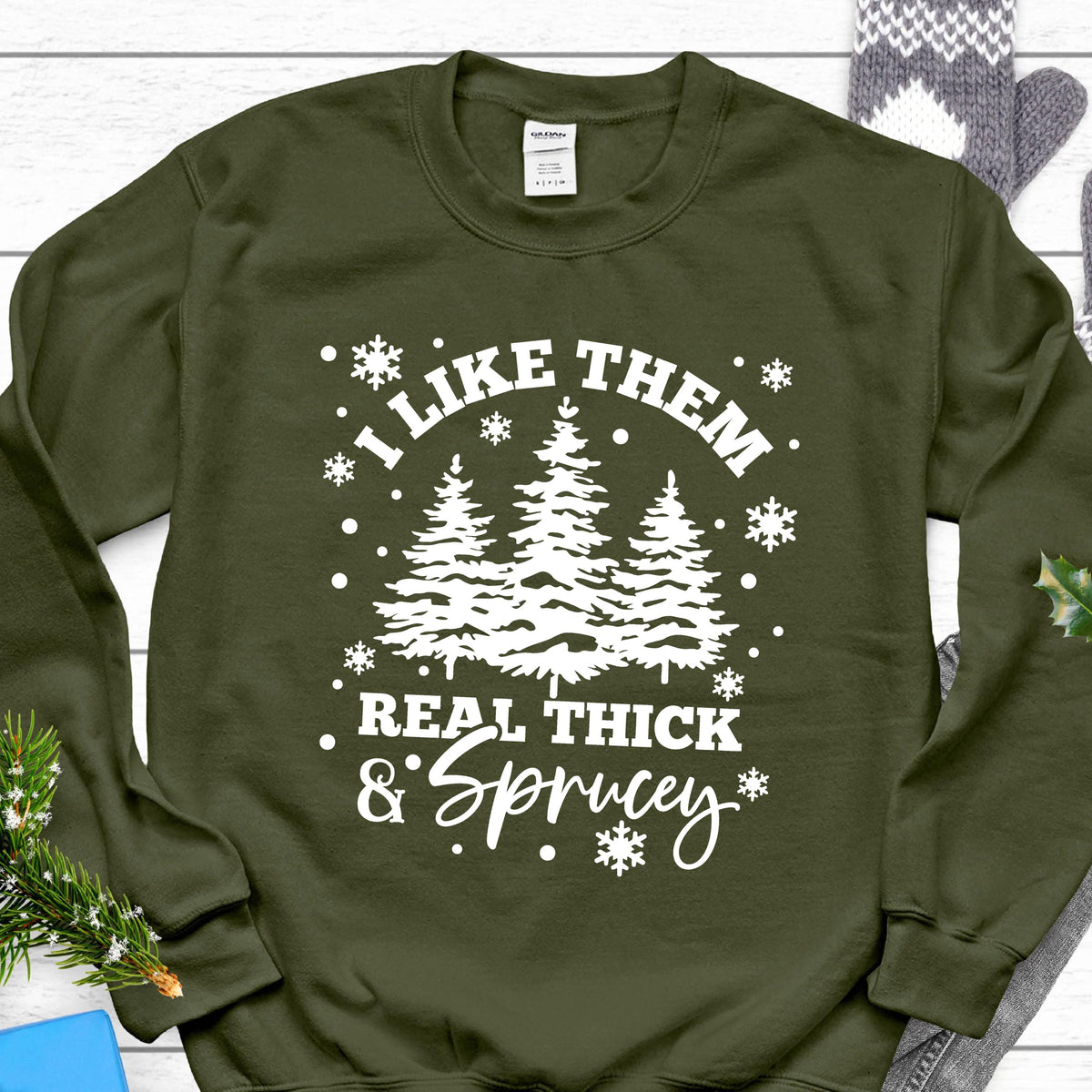 May include: An olive green crewneck sweatshirt featuring a white graphic print of three pine trees surrounded by snowflakes. The text reads, "I LIKE THEM REAL THICK & SPRUCEY" in a combination of bold block lettering and a cursive script font.