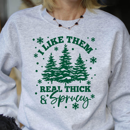 May include: A light gray heather crewneck sweatshirt featuring a dark green graphic print of three pine trees, snowflakes, and the text "I LIKE THEM REAL THICK & SPRUCEY" in a combination of bold block lettering and a cursive script font.