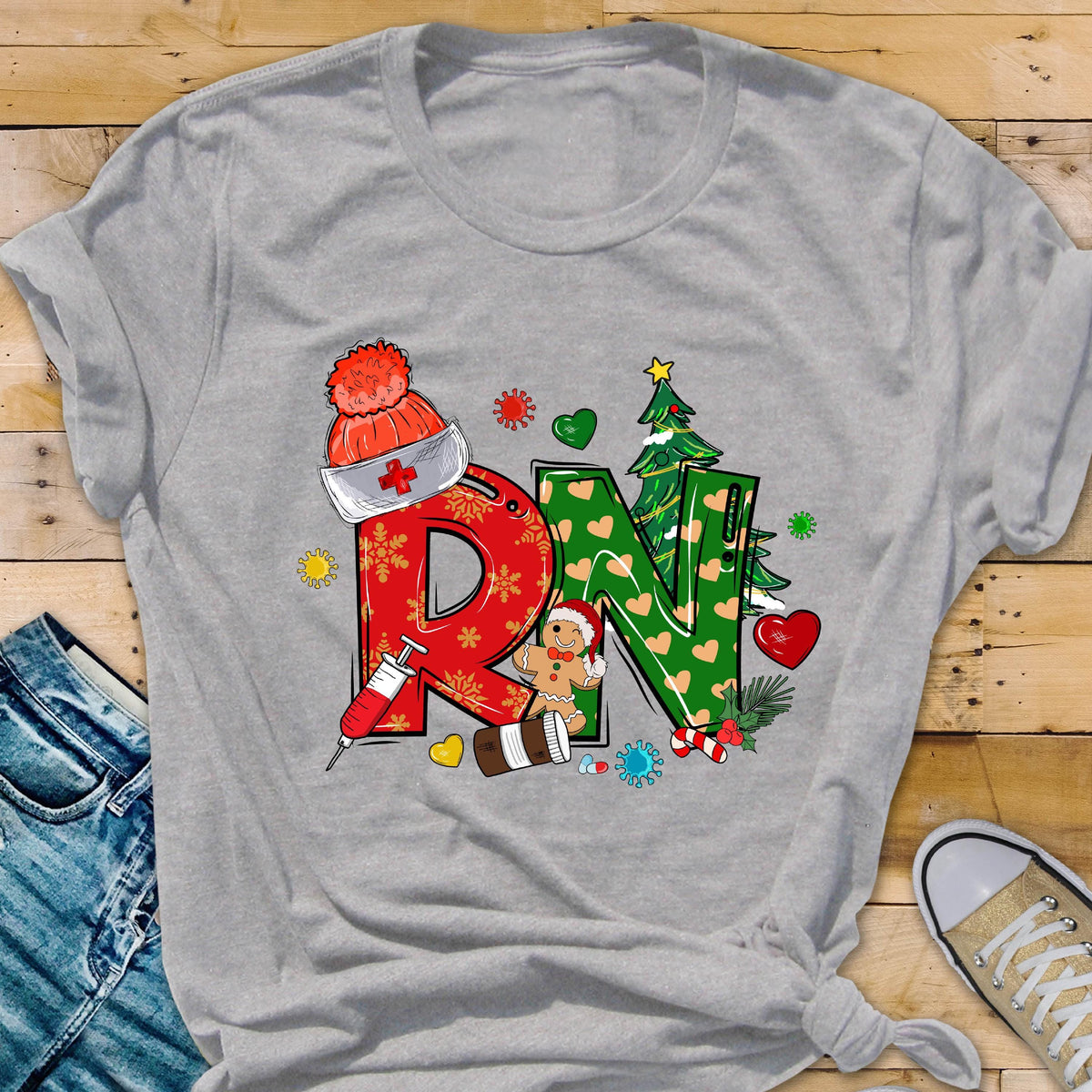 May include: A heather gray t-shirt featuring a festive design for nurses. The design includes the letters "RN" decorated with Christmas-themed elements like a Santa hat, gingerbread man, Christmas tree, and syringe.