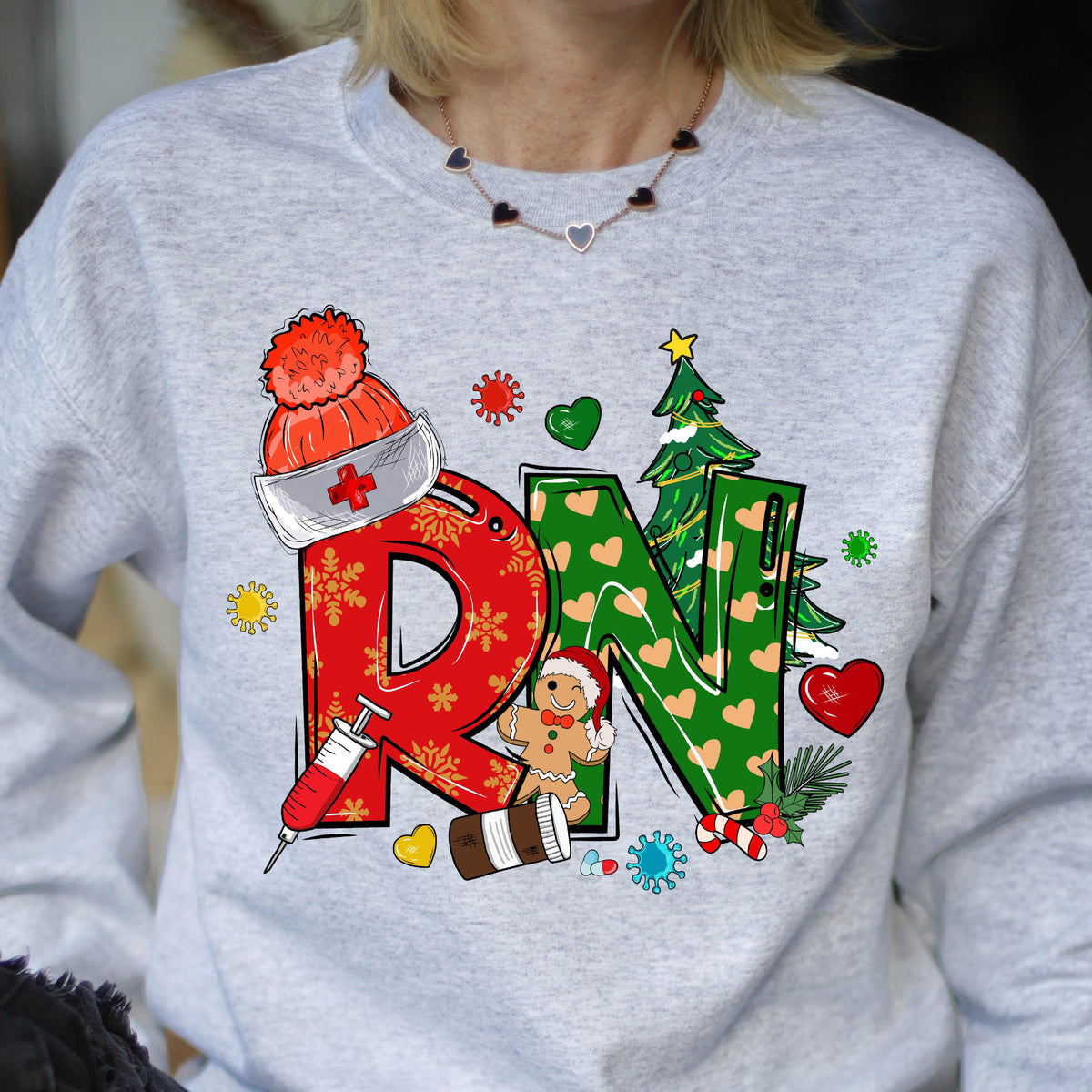 May include: A light gray crewneck sweatshirt featuring a large red and green graphic with the letters RN. The design includes a red nurse cap, a syringe, a gingerbread person, a Christmas tree, hearts, and various colorful virus-like shapes.