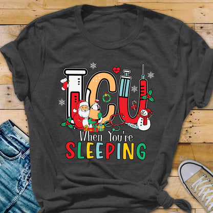 May include: Dark gray t-shirt with a colorful graphic design. The design features the letters "ICU" in orange and red, along with a Santa Claus, a snowman, and medical equipment. The text "When You're SLEEPING" is below the graphic.