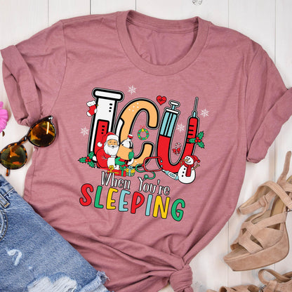 May include: A mauve-colored t-shirt featuring a colorful graphic design. The design includes the letters "ICU" in a festive font, along with images of Santa Claus, a snowman, and medical equipment. The text "When You're Sleeping" is also included.