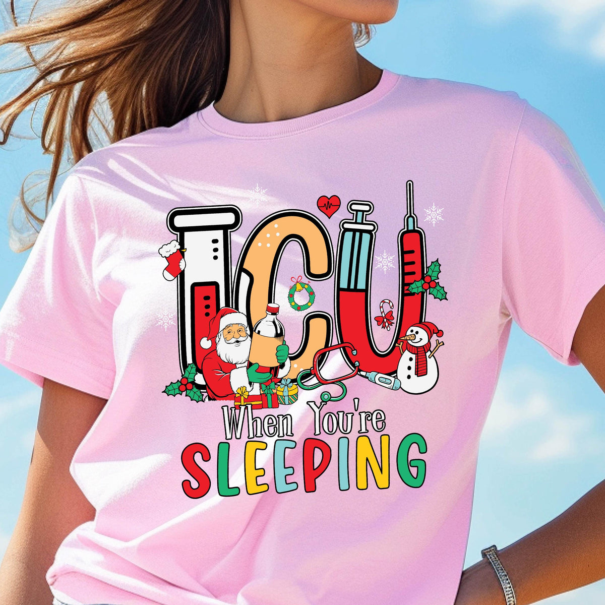 May include: A light pink t-shirt with a colorful graphic design. The design features the letters "ICU" in black and orange, with medical and Christmas-themed illustrations. The text "When You're Sleeping" is below the letters in green and orange.