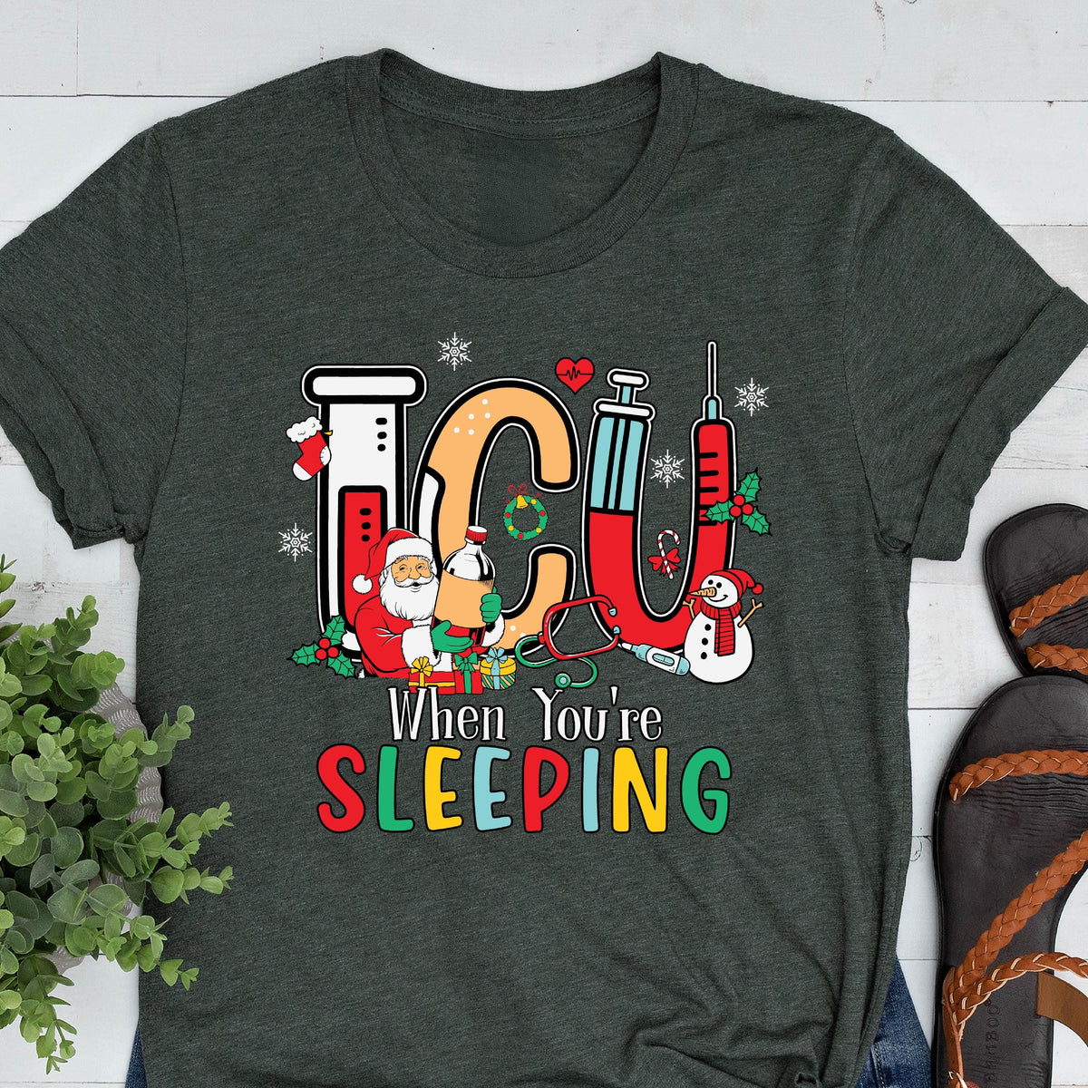 May include: Dark gray t-shirt with a colorful graphic design. The design features the letters "ICU" in red and orange, with medical and Christmas-themed elements. Text below reads "When You're SLEEPING."