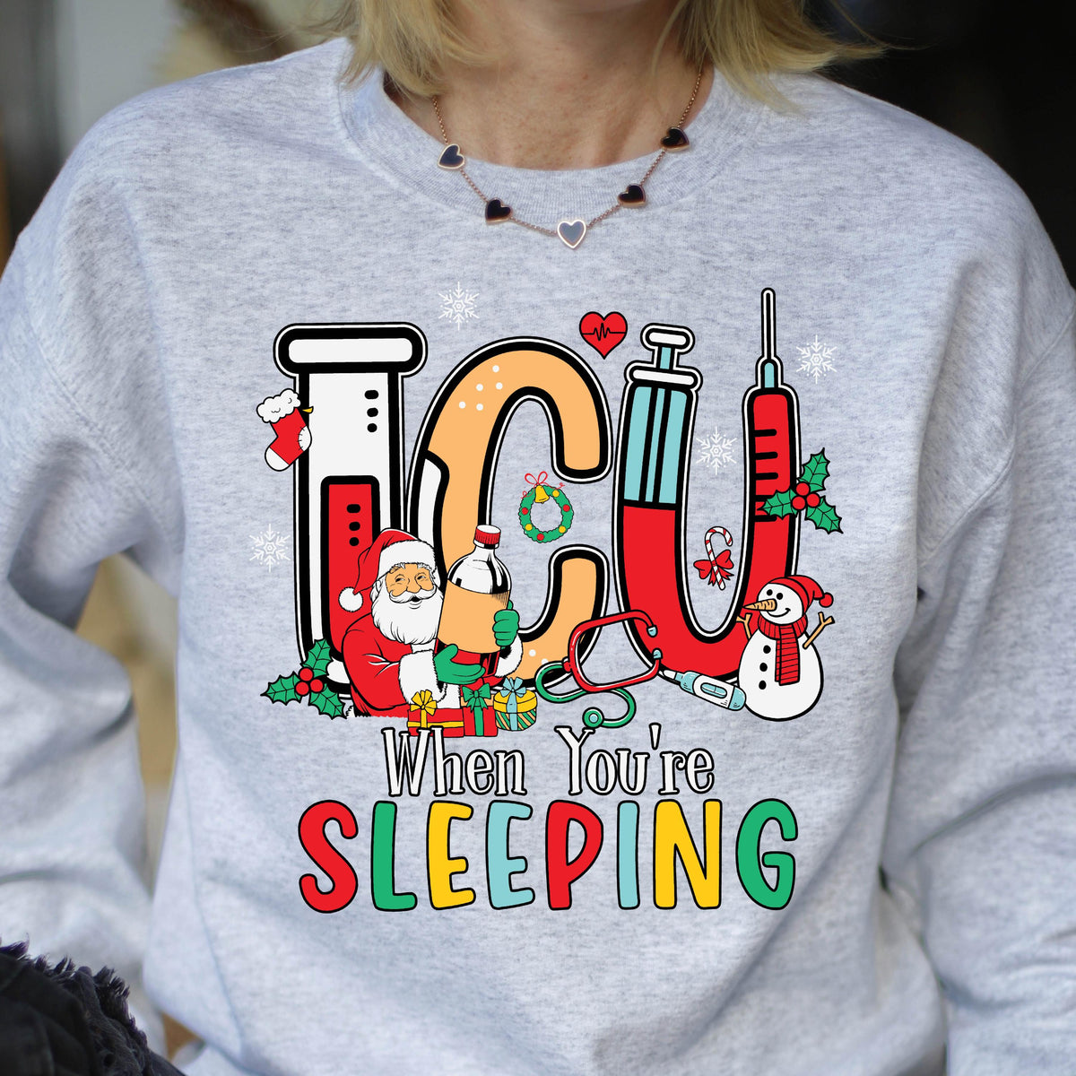 May include: A light gray sweatshirt with a colorful graphic design. The design features the letters "ICU" in orange and red, with medical and Christmas-themed illustrations. Text below reads "When You're SLEEPING."