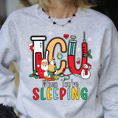 May include: A light gray sweatshirt with a colorful graphic design. The design features the letters "ICU" in orange and red, with medical and Christmas-themed illustrations. Text below reads "When You're SLEEPING."