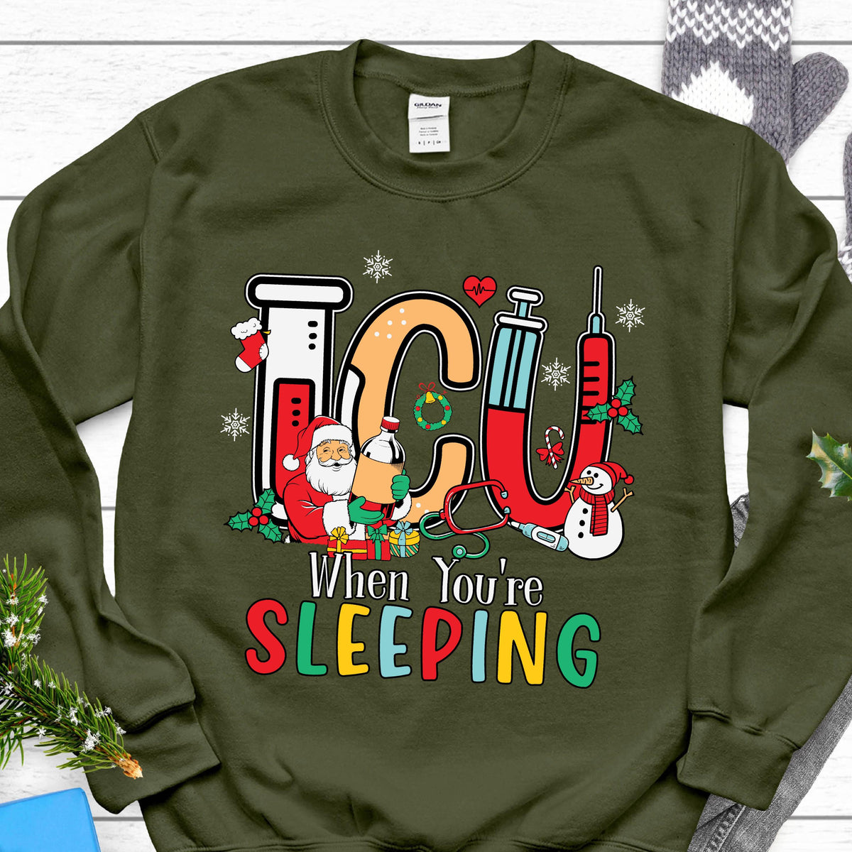 May include: Green sweatshirt with a festive ICU design. The design includes the letters "ICU" in a medical theme with a Santa Claus, snowman, and the text "When You're Sleeping" in colorful letters. A Christmas-themed gift.