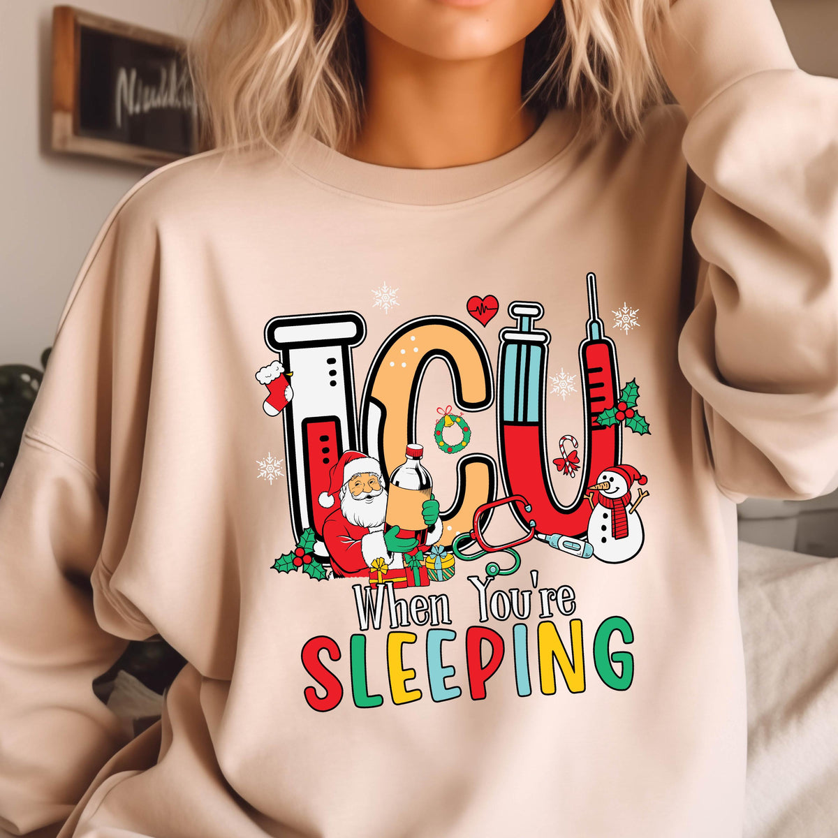 May include: Beige sweatshirt with a colorful graphic. The graphic spells "ICU" with medical and Christmas-themed illustrations. Santa Claus, a snowman, and a syringe are featured. The text "When You're Sleeping" is below the ICU letters.
