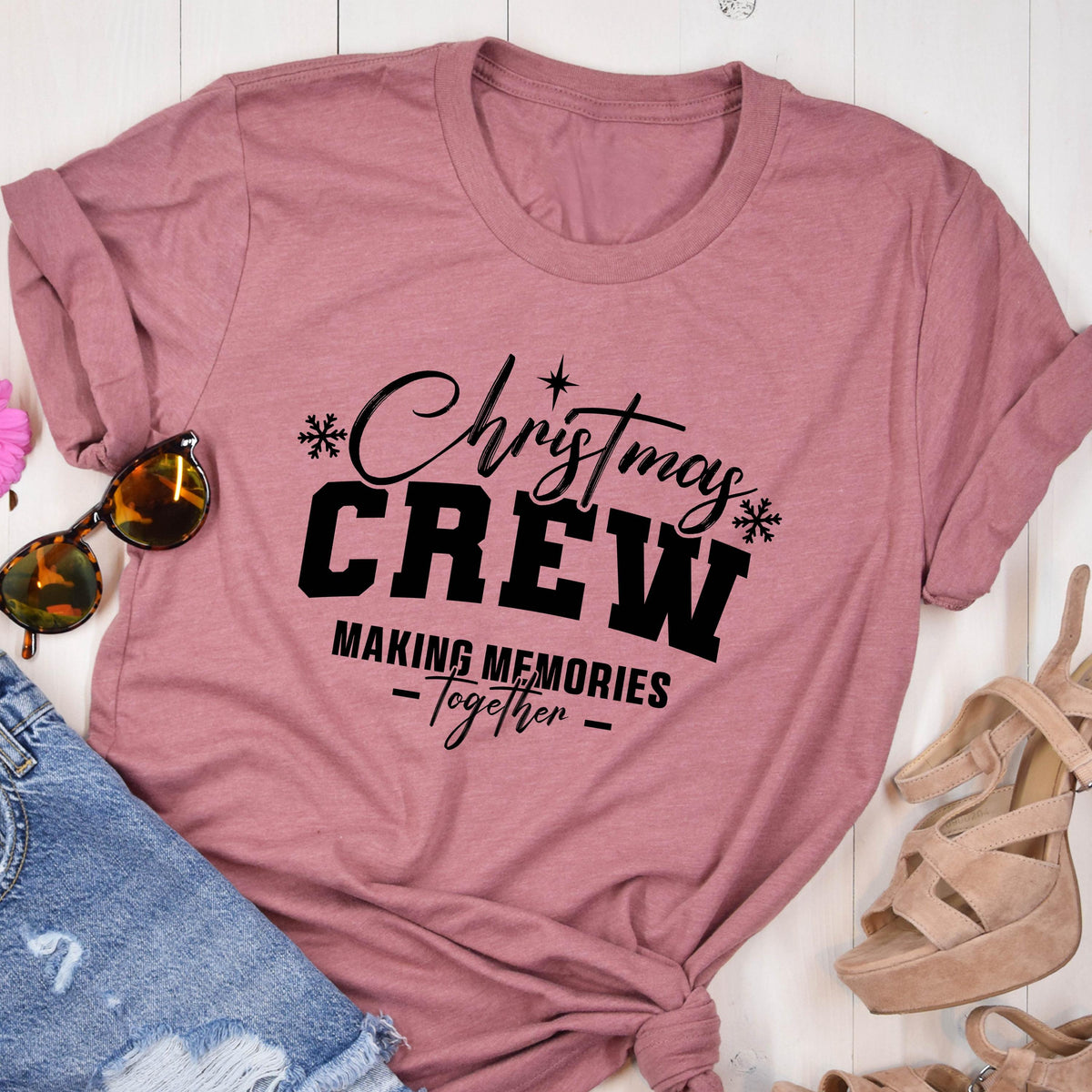 May include: A mauve short-sleeved t-shirt featuring black text that reads Christmas CREW MAKING MEMORIES Together, accented with small snowflake and star graphics. The shirt is displayed alongside sunglasses, denim fabric, and beige heels.