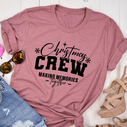 May include: A mauve short-sleeved t-shirt featuring black text that reads Christmas CREW MAKING MEMORIES Together, accented with small snowflake and star graphics. The shirt is displayed alongside sunglasses, denim fabric, and beige heels.