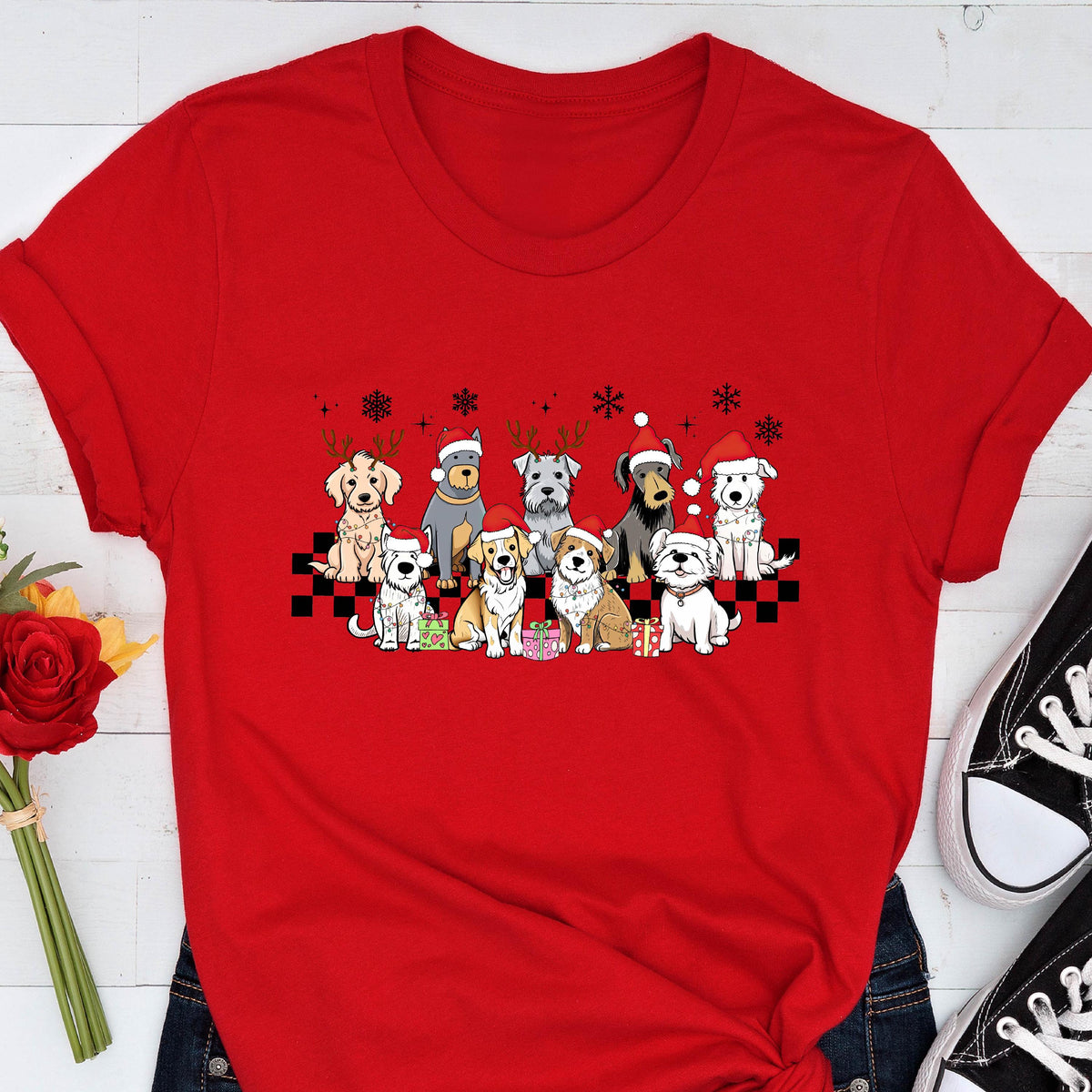 May include: Red t-shirt featuring a graphic of various dog breeds wearing Santa hats and reindeer antlers. The dogs are arranged on a black and white checkered pattern with snowflakes. A festive holiday-themed apparel item.