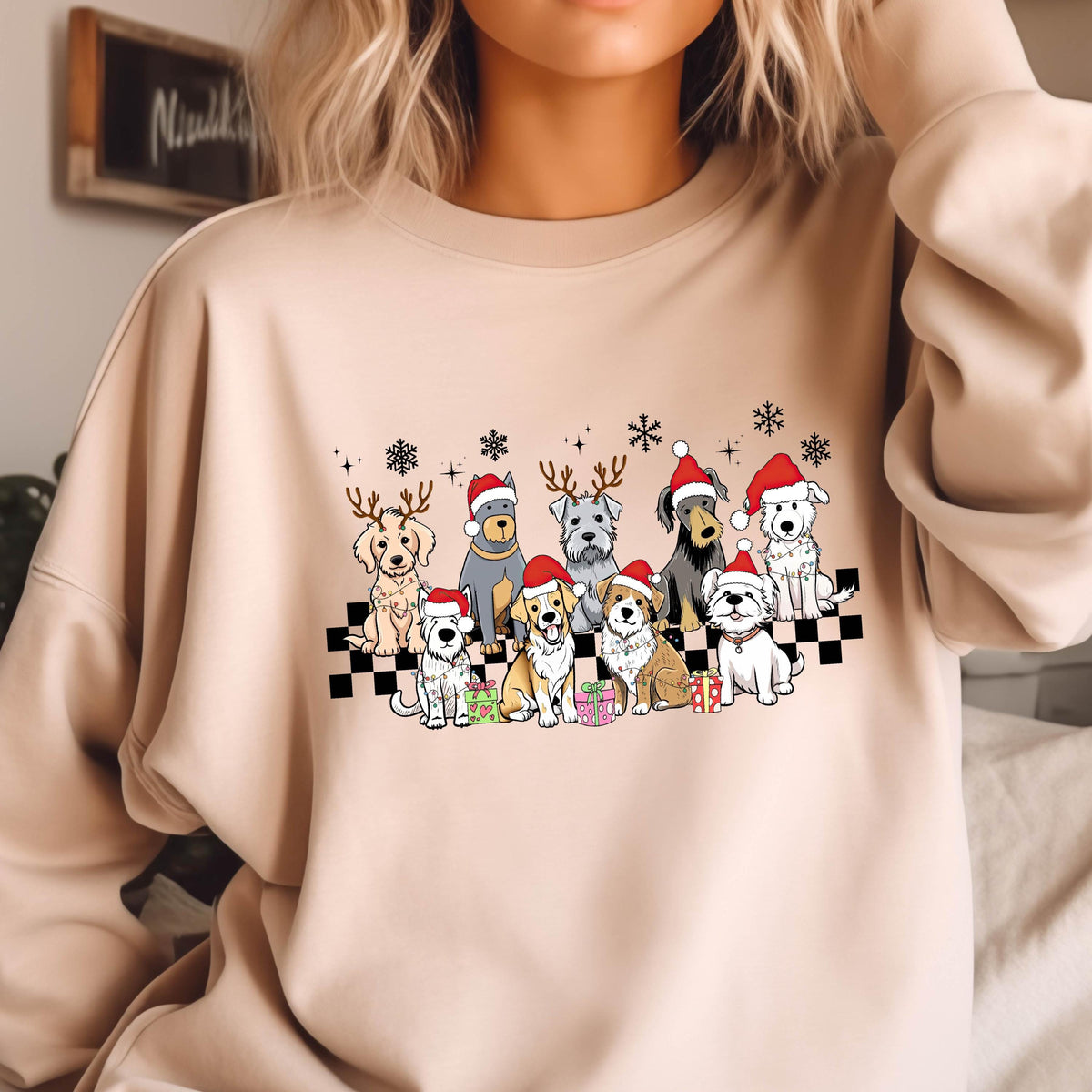 May include: Beige sweatshirt featuring a festive graphic of various dog breeds wearing Santa hats and reindeer antlers. The dogs are arranged on a black and white checkered pattern, with snowflakes and gift boxes. A cozy and cheerful design.