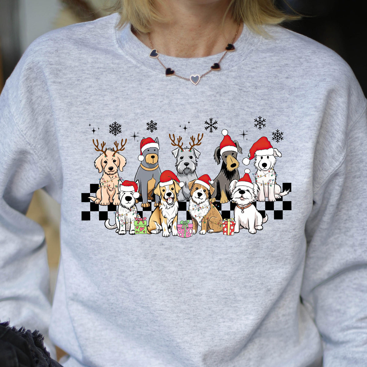 May include: A light gray sweatshirt featuring a festive design of various dog breeds wearing Santa hats and reindeer antlers. The dogs are arranged on a black and white checkered pattern, with snowflakes above and wrapped gifts below.