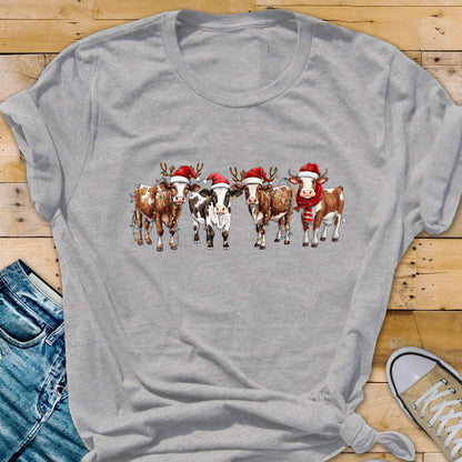 May include: A heather gray t-shirt featuring a festive design of five cows. Each cow is adorned with a Santa hat and some with reindeer antlers and Christmas lights. One cow wears a red scarf. The cows are brown, white, and black.