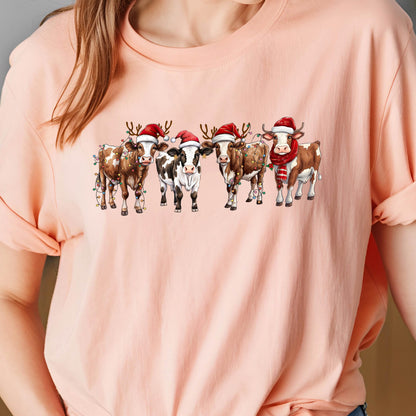 May include: A light pink t-shirt featuring a festive design of five cows. Each cow is adorned with a Santa hat, antlers, and some are wearing a scarf. The cows are decorated with Christmas lights, creating a holiday theme.