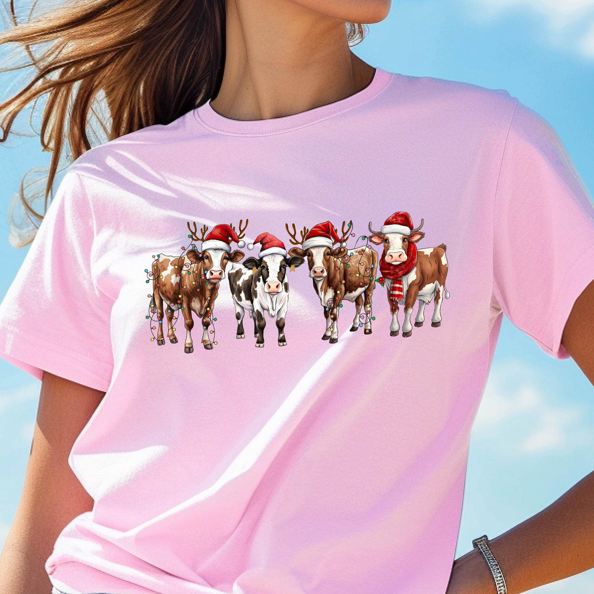 May include: A light pink t-shirt featuring a festive design of five cows. Each cow is adorned with a Santa hat, and some have reindeer antlers and Christmas lights. One cow wears a red and white striped scarf.