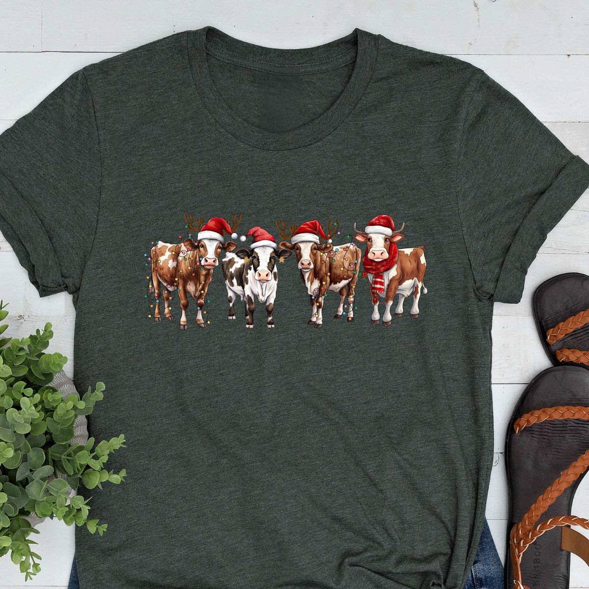 May include: Dark gray t-shirt featuring a festive design of five cows. Each cow is adorned with a Santa hat and some have reindeer antlers and scarves. The cows are brown and white, creating a holiday-themed graphic.