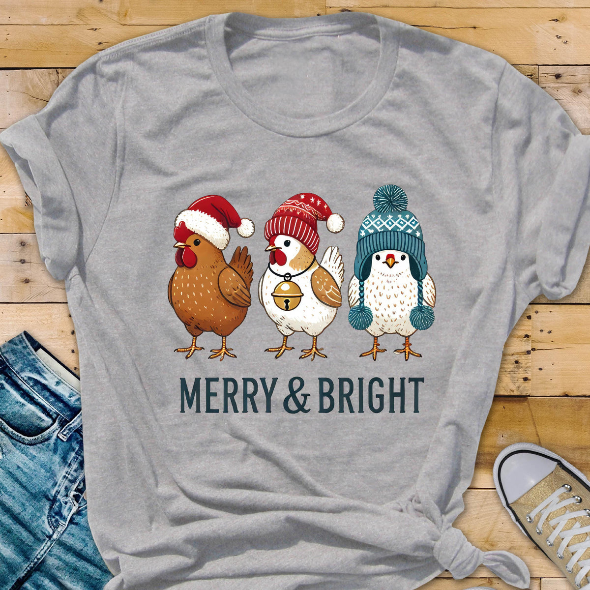 May include: A heather gray t-shirt featuring three cartoon chickens wearing festive winter hats. The chickens are brown, white, and speckled, with a bell on one. The text "MERRY & BRIGHT" is printed below the chickens.
