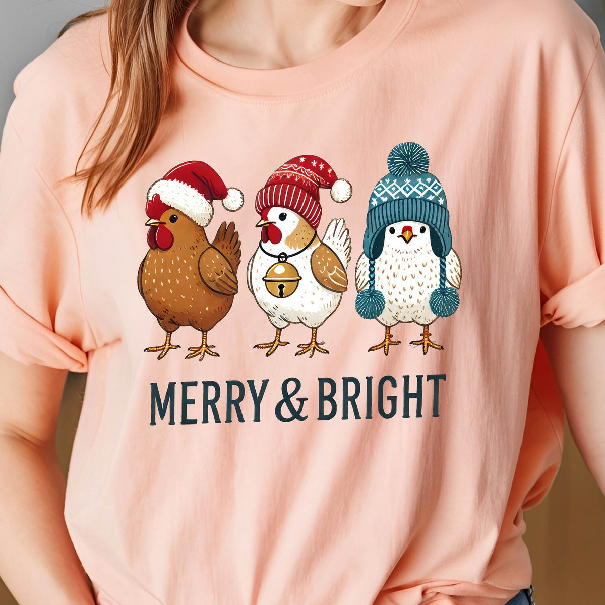 May include: A light pink t-shirt featuring a graphic of three chickens wearing festive hats. The chickens are brown, white, and white with a blue hat. The text "MERRY & BRIGHT" is printed below the chickens in navy blue.