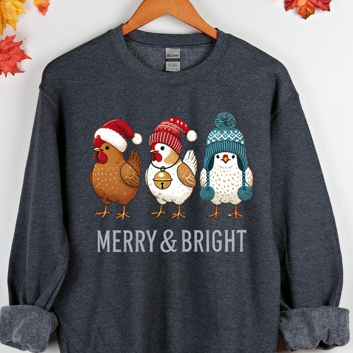 May include: A dark gray sweatshirt with a Christmas-themed design featuring three chickens wearing Santa hats and winter beanies. The text "Merry & Bright" is printed below the chickens.