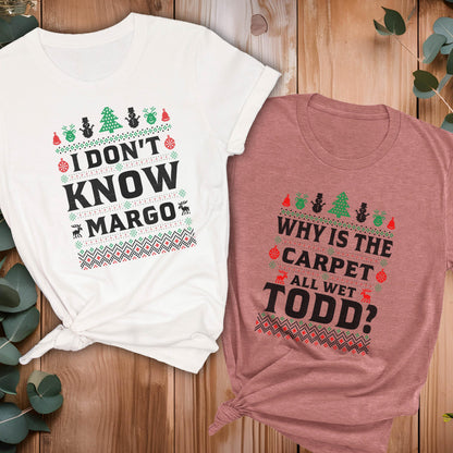May include: Two short-sleeved t-shirts with Christmas-themed designs. One is white with the text "I DON'T KNOW MARGO" and the other is mauve with the text "WHY IS THE CARPET ALL WET TODD?". Both shirts have festive graphics.