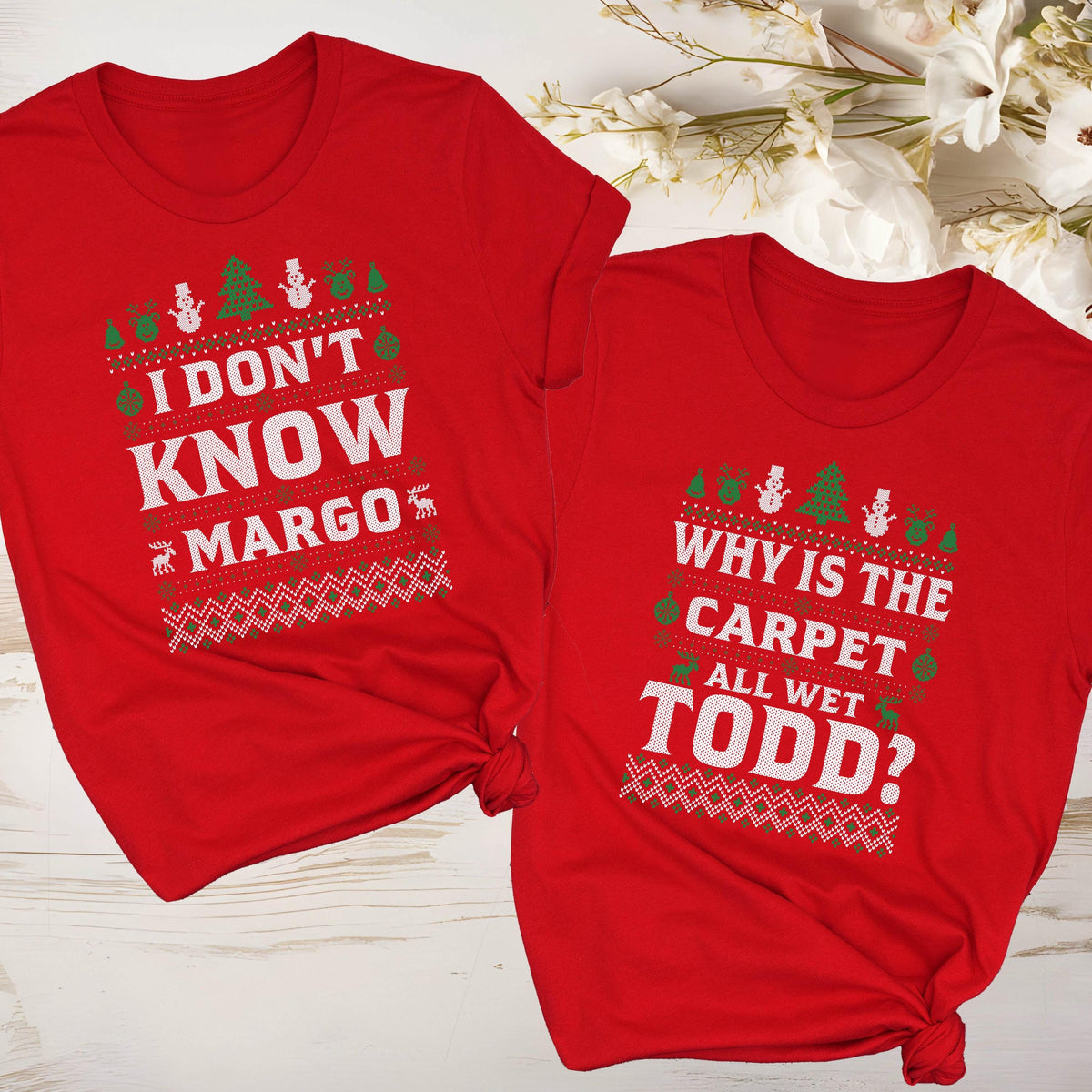 May include: Two red t-shirts with holiday-themed text. One shirt says "I DON'T KNOW MARGO" with festive graphics. The other says "WHY IS THE CARPET ALL WET TODD?" with similar designs. Both shirts have a crew neck.