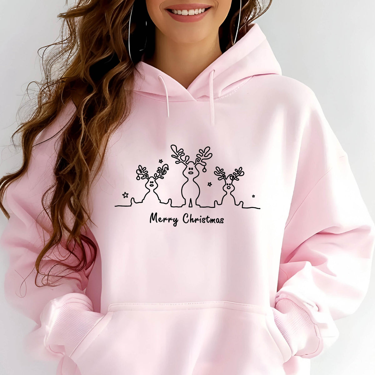 May include: A light pink hooded sweatshirt featuring a black line art graphic of three reindeer with antlers and small stars. Below the reindeer, the text Merry Christmas is printed in a black script font. The hoodie includes a front kangaroo pocket.